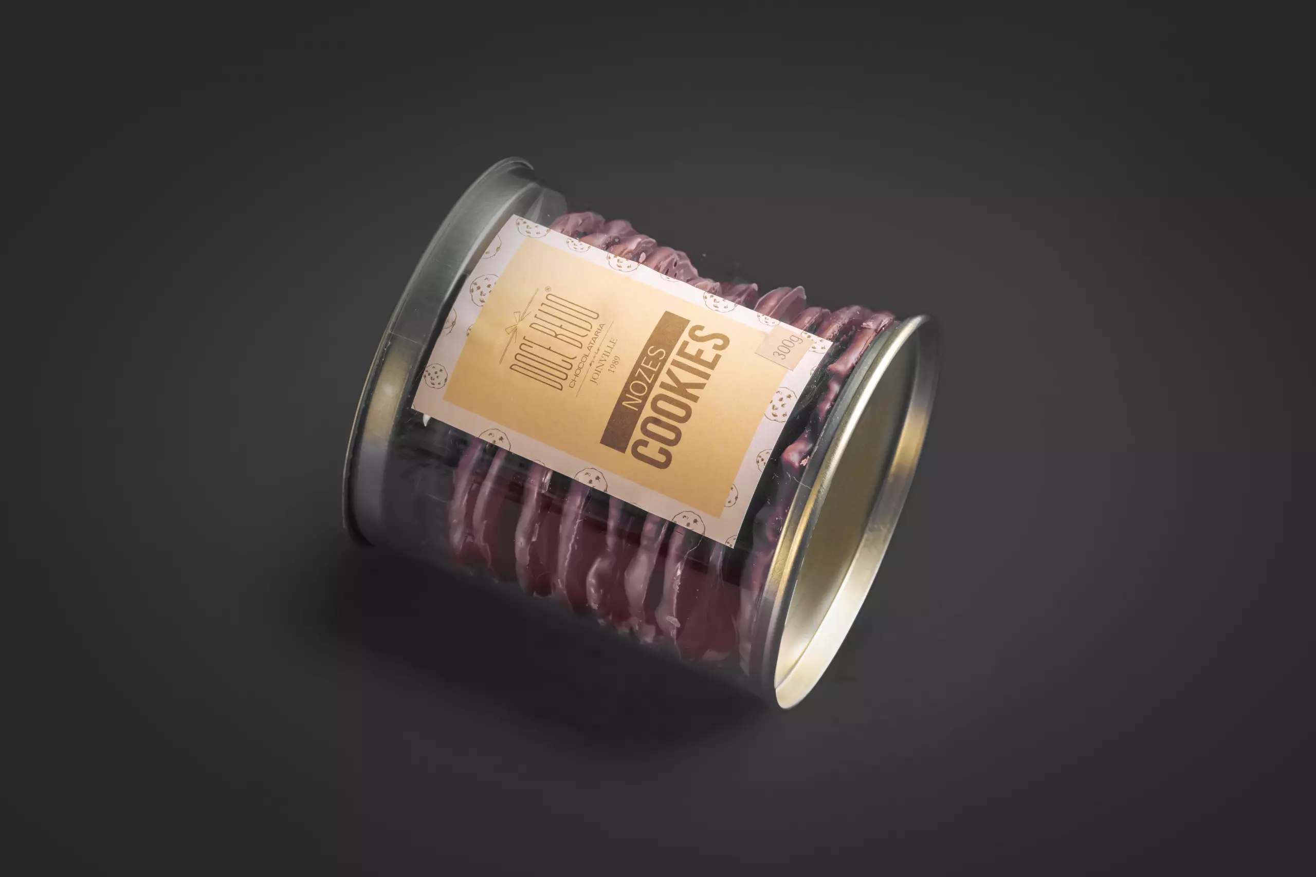 Um pote cilíndrico de plástico transparente, com tampa e base prateadas, deitado de lado e cheio de cookies de nozes empilhados. Um rótulo de papel bege na lateral identifica o produto como "NOZES COOKIES" da marca "Doce Beijo". O fundo é cinza-escuro e uniforme.