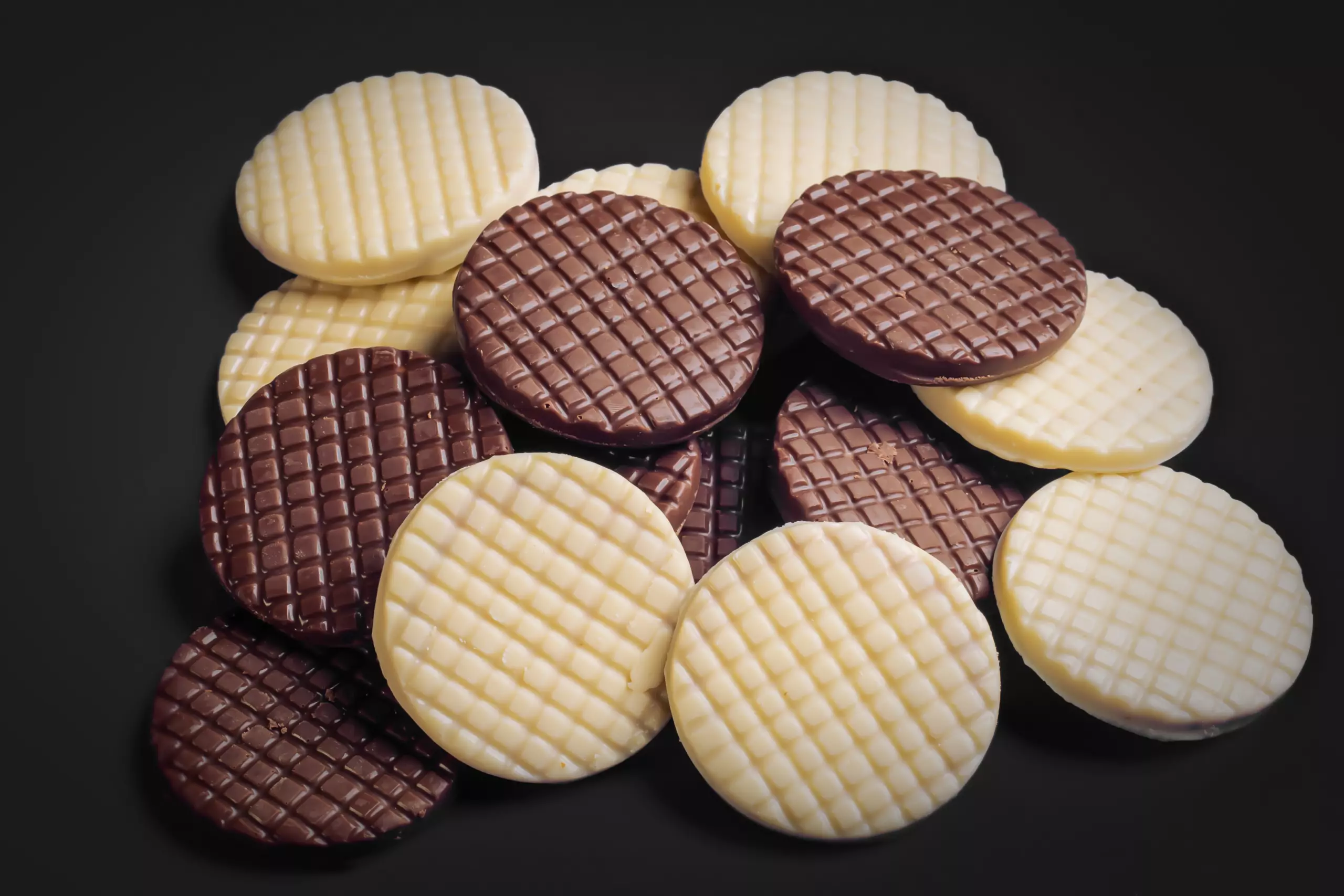Uma pilha de biscoitos redondos do tipo "Sweet Salty" sobre um fundo preto. Os biscoitos estão misturados, sendo alguns cobertos com chocolate branco e outros com chocolate amargo, e todos possuem uma textura quadriculada na superfície.