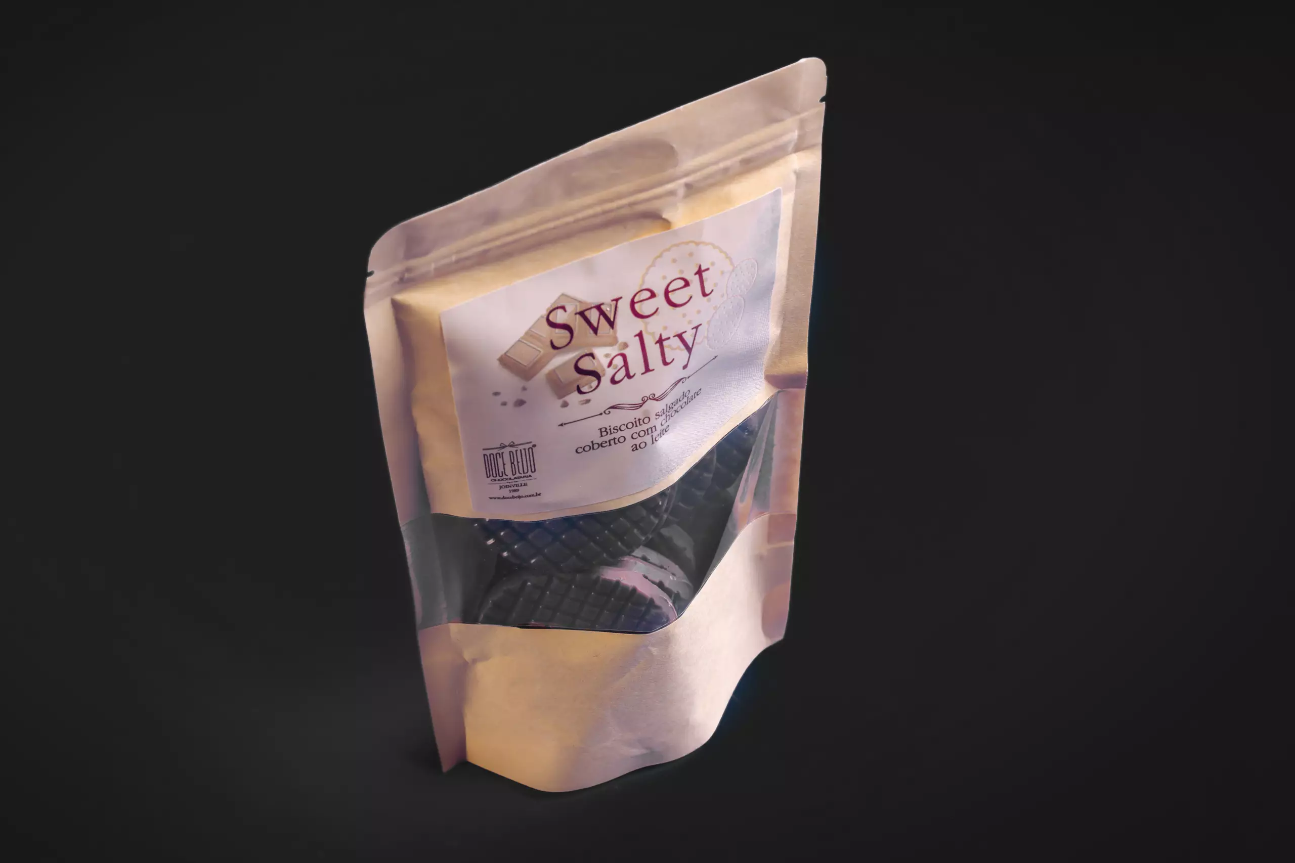 Uma embalagem do tipo "stand-up pouch" do produto "Sweet Salty". O pacote de papel kraft tem uma janela transparente que mostra os biscoitos redondos, nos sabores chocolate ao leite em seu interior. O rótulo informa que se trata de um "Biscoito salgado coberto com chocolate ao leite".