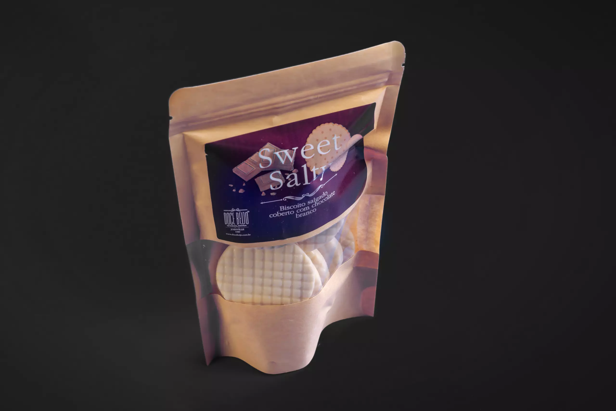Uma embalagem do tipo "stand-up pouch" do produto "Sweet Salty". O rótulo descreve o conteúdo como "Biscoito salgado coberto com chocolate branco". Através da janela da embalagem, são visíveis os biscoitos redondos e brancos com textura quadriculada.