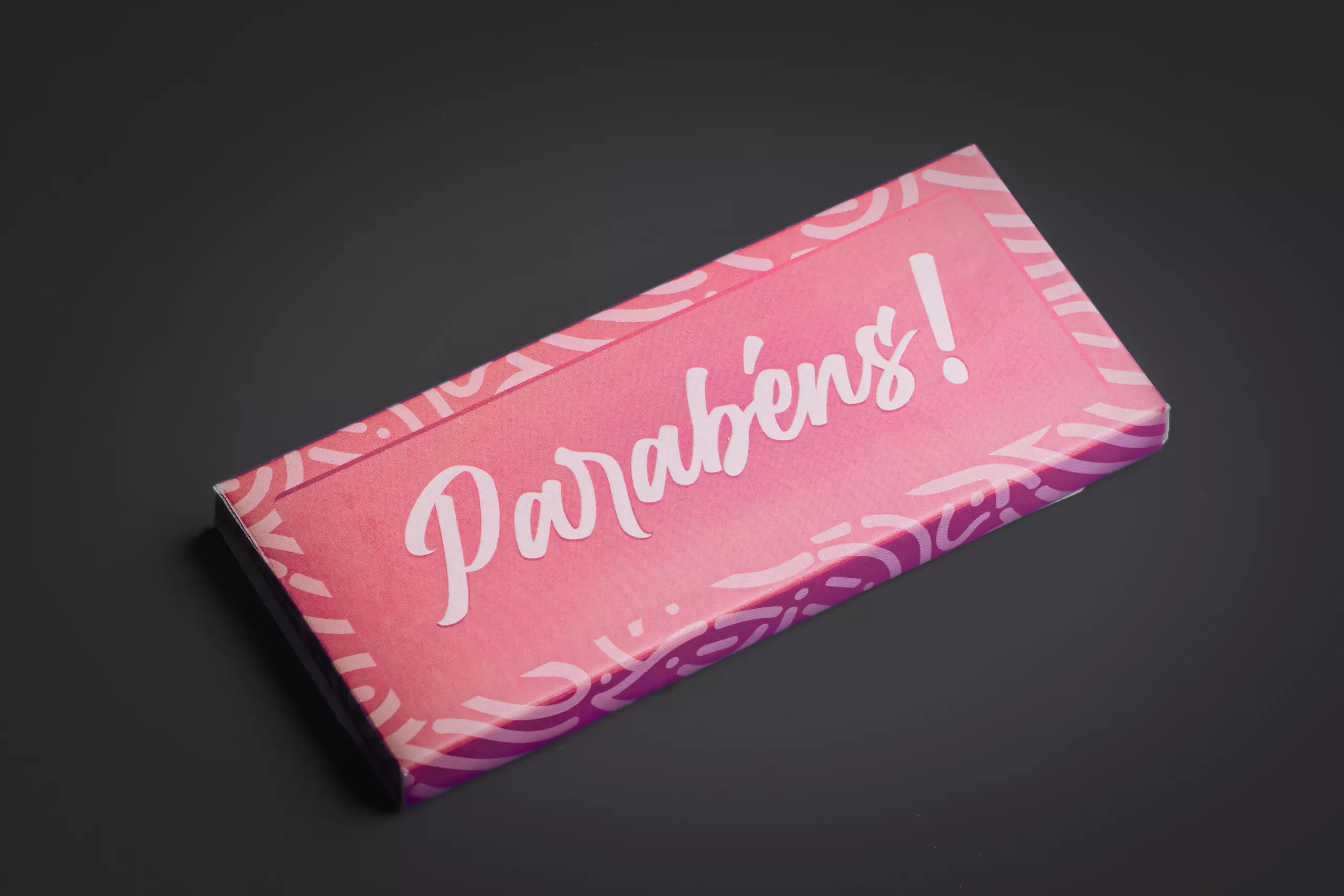Uma barra de chocolate retangular com uma embalagem de papel na cor rosa. A palavra "Parabéns!" está escrita em branco no centro, com uma fonte grande e estilizada. A barra está disposta em ângulo sobre um fundo cinza-escuro.