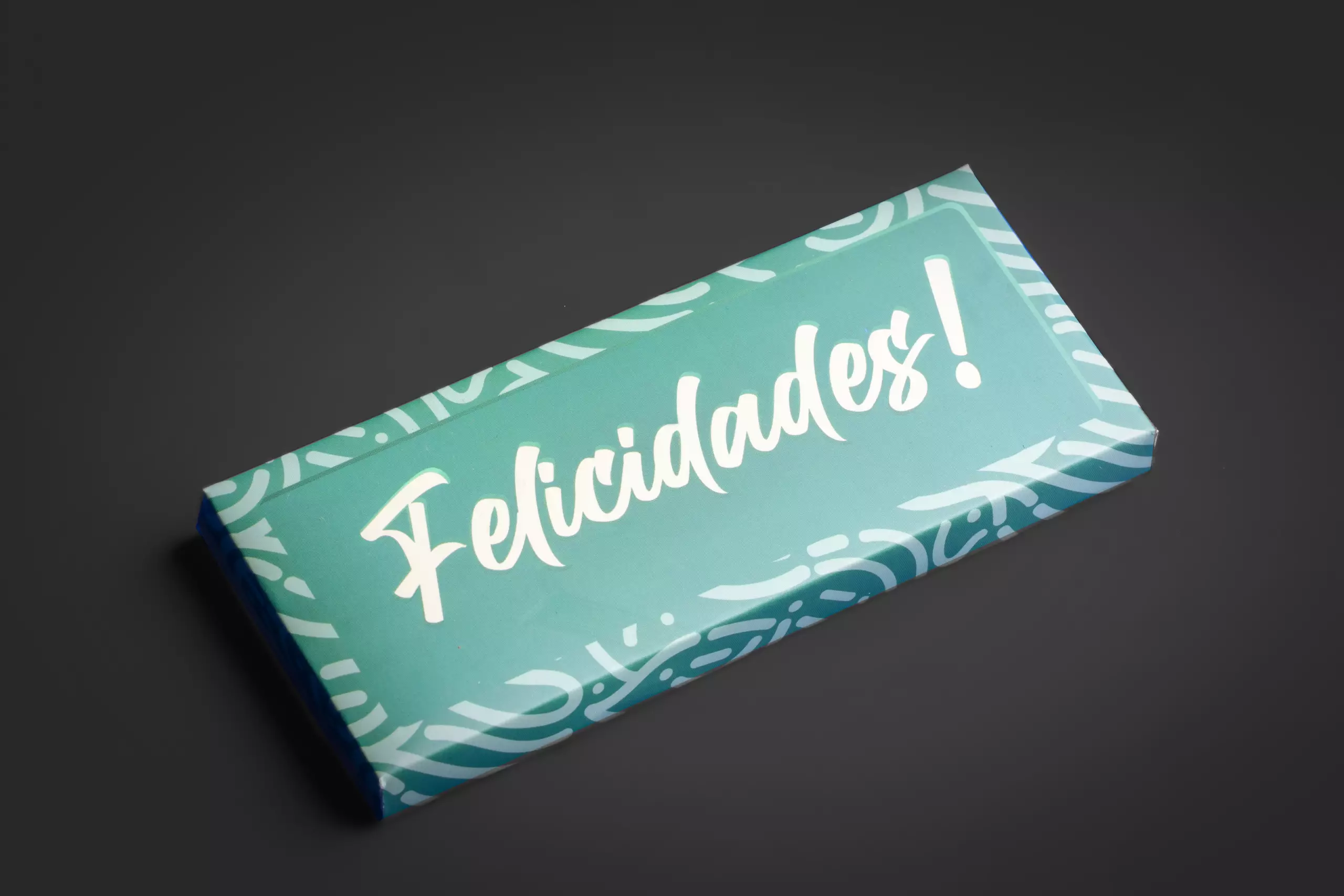Uma barra de chocolate retangular com uma embalagem decorativa na cor verde-água. A palavra "Felicidades!" está escrita em branco no centro, com uma fonte grande e estilizada. A barra está disposta em ângulo sobre um fundo cinza-escuro uniforme.