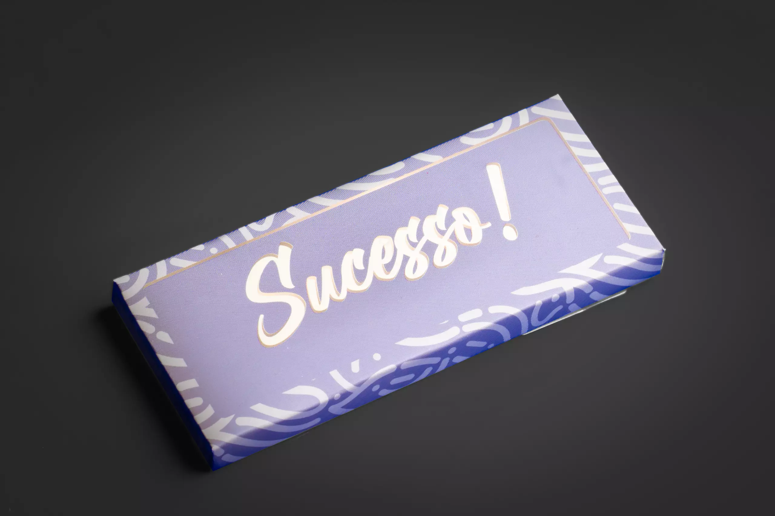 Uma barra de chocolate retangular com uma embalagem de papel na cor lilás. A palavra "Sucesso!" está escrita em branco e dourado no centro, com uma fonte grande e estilizada. A barra está sobre um fundo cinza-escuro uniforme.