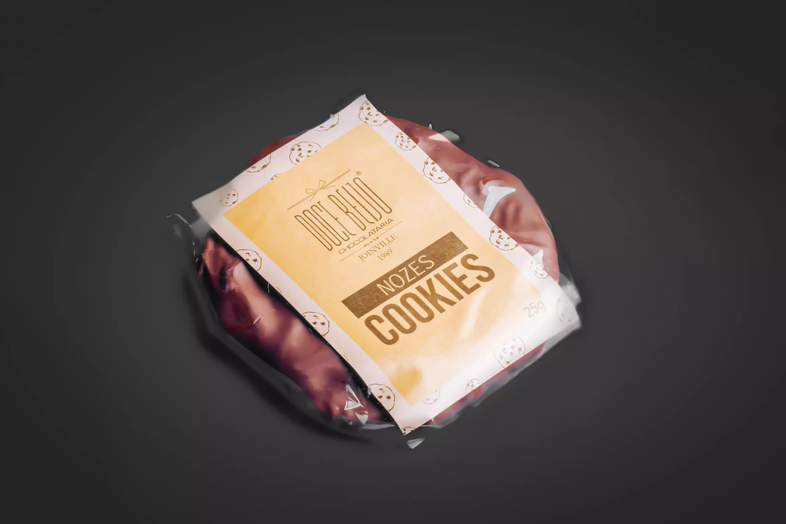 Um pacote plástico transparente, fotografado de cima, contendo um cookies redondo. Uma etiqueta bege e branca, com o logotipo "Doce Beijo" e a inscrição "NOZES COOKIES", está fixada sobre a embalagem. Pequenos desenhos de cookies adornam a etiqueta. O fundo é escuro e uniforme.