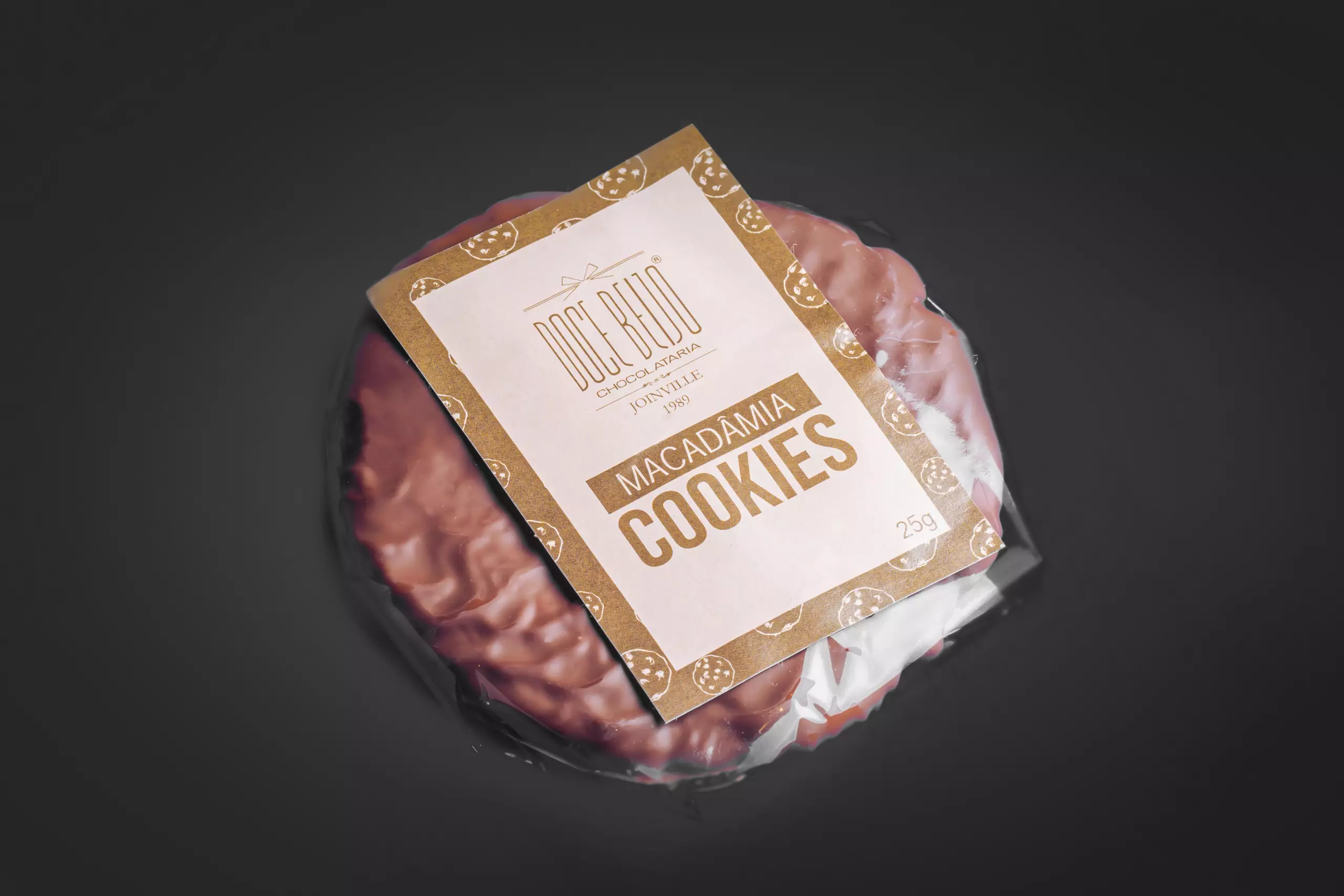 Um pacote plástico transparente, visto de cima, contendo um cookies redondos. Uma etiqueta bege e branca, com o logotipo "Doce Beijo" e a inscrição "MACADÂMIA COOKIES", está colada sobre a embalagem. Pequenos desenhos de cookies decoram a etiqueta. O fundo é escuro e uniforme.