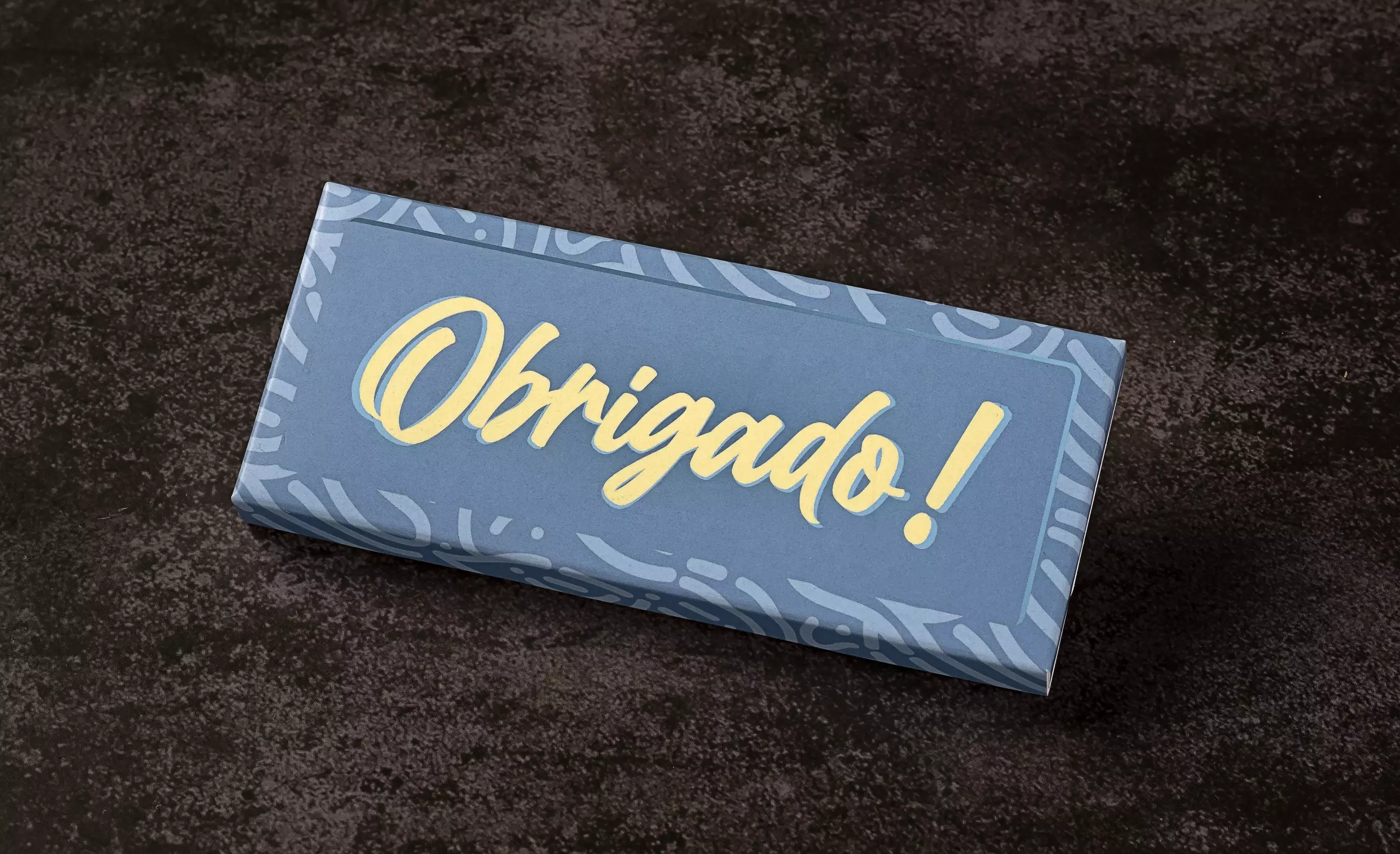 Uma barra de chocolate retangular com uma embalagem de papel na cor azul-acinzentado. A palavra "Obrigado!" está escrita em amarelo no centro, com uma fonte grande e estilizada. A barra está sobre uma superfície escura e texturizada.