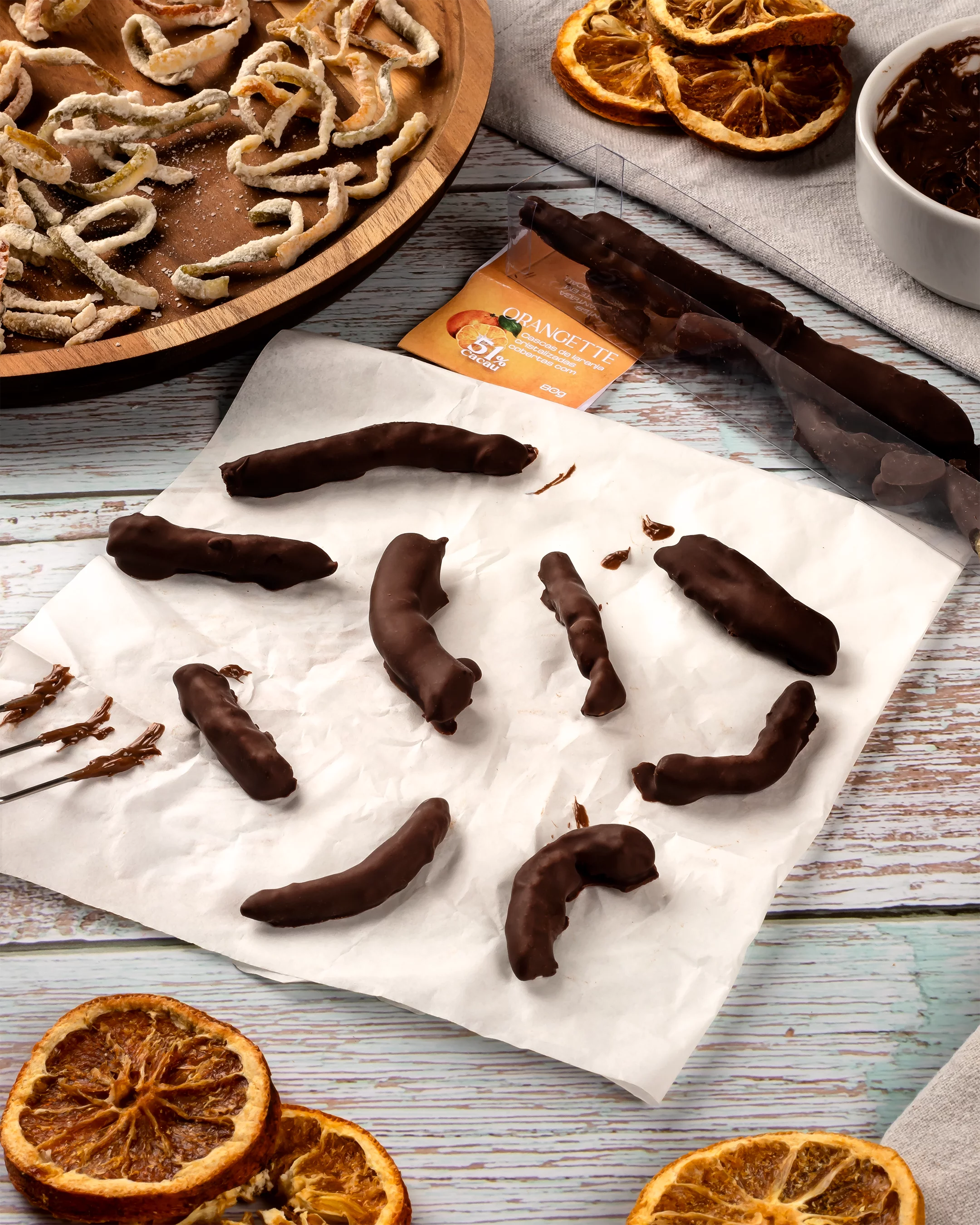Orangettes feitos de tiras de casca de laranja cristalizadas sendo banhadas em chocolate amargo e dispostas sobre um papel manteiga para secar. Ao redor, há um prato de madeira com mais cascas de laranja, rodelas de laranja desidratadas, um pote com chocolate derretido e a embalagem do produto "Orangettes 51% cacau".