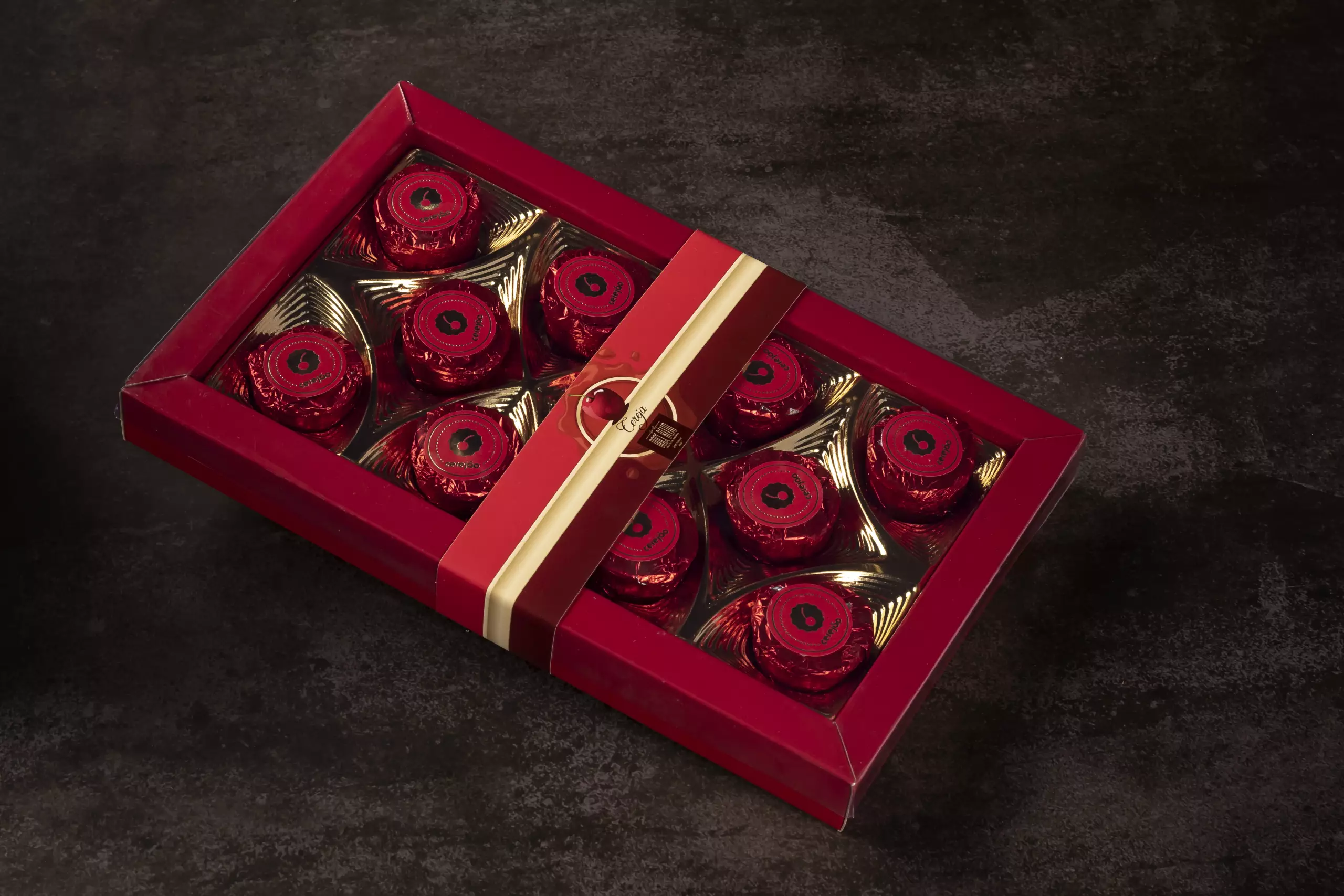 Uma caixa de presente retangular e vermelha, aberta, contendo dez bombons "Cerejão". Cada bombom é embalado individualmente em papel laminado vermelho e está acomodado em um suporte dourado. A caixa está disposta sobre uma superfície escura e texturizada.