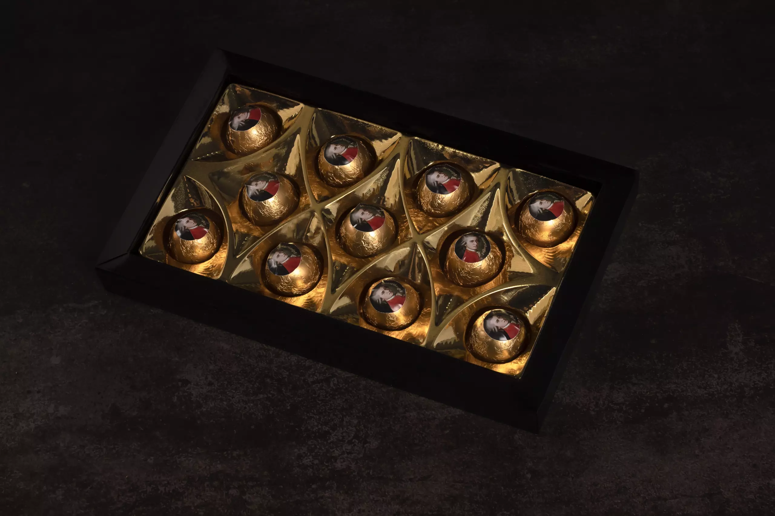 Caixa preta contendo 12 bombons Mozartkugel embalados em papel dourado, cada um com selo no topo estampando o retrato de Mozart. A caixa está aberta, revelando o interior dourado e o arranjo elegante dos bombons, disposta sobre uma superfície escura.