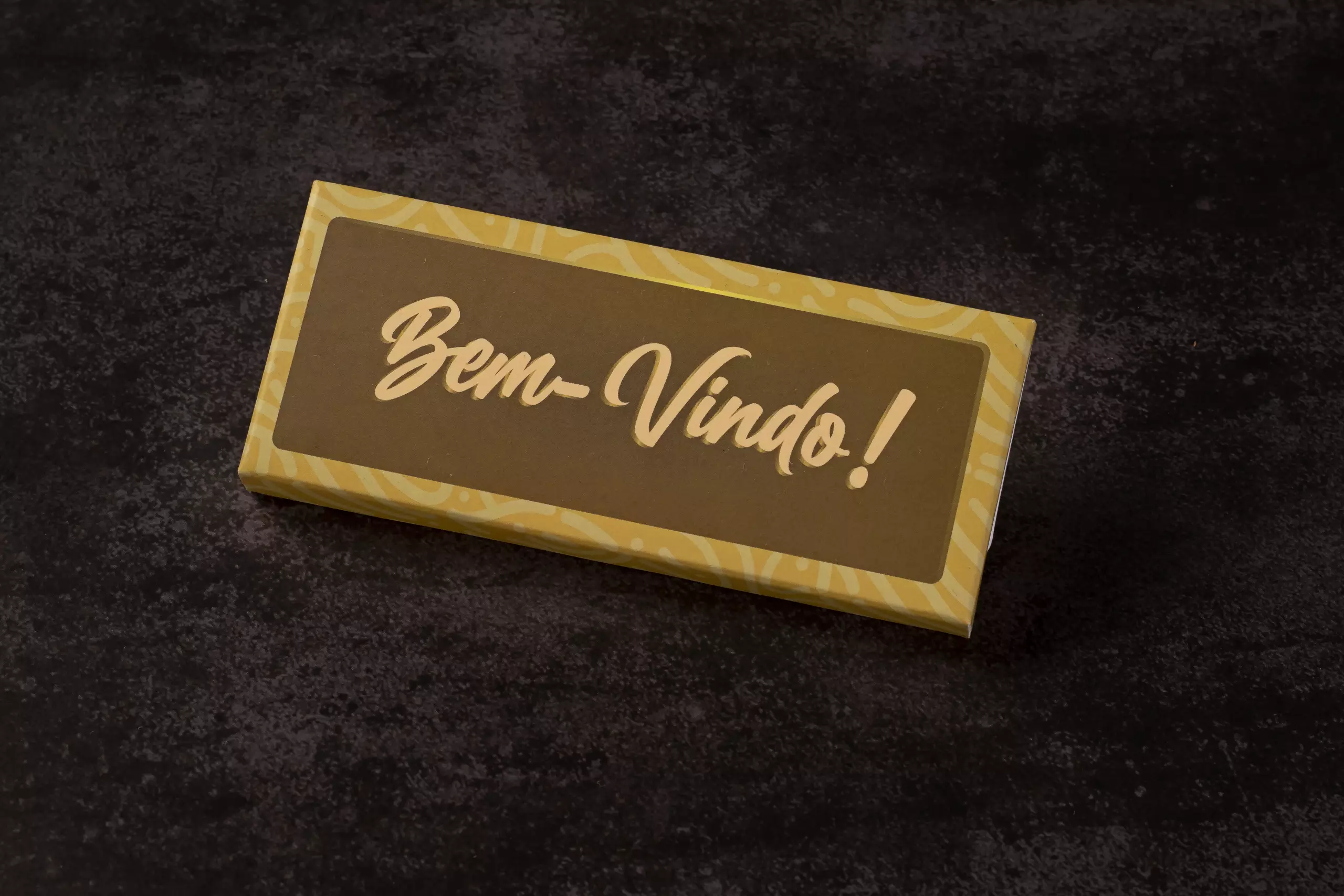 Uma barra de chocolate retangular com uma embalagem de papel em tons de marrom e dourado. A frase "Bem-Vindo!" está escrita no centro com uma fonte amarela e estilizada. A barra está disposta em ângulo sobre uma superfície escura e texturizada.