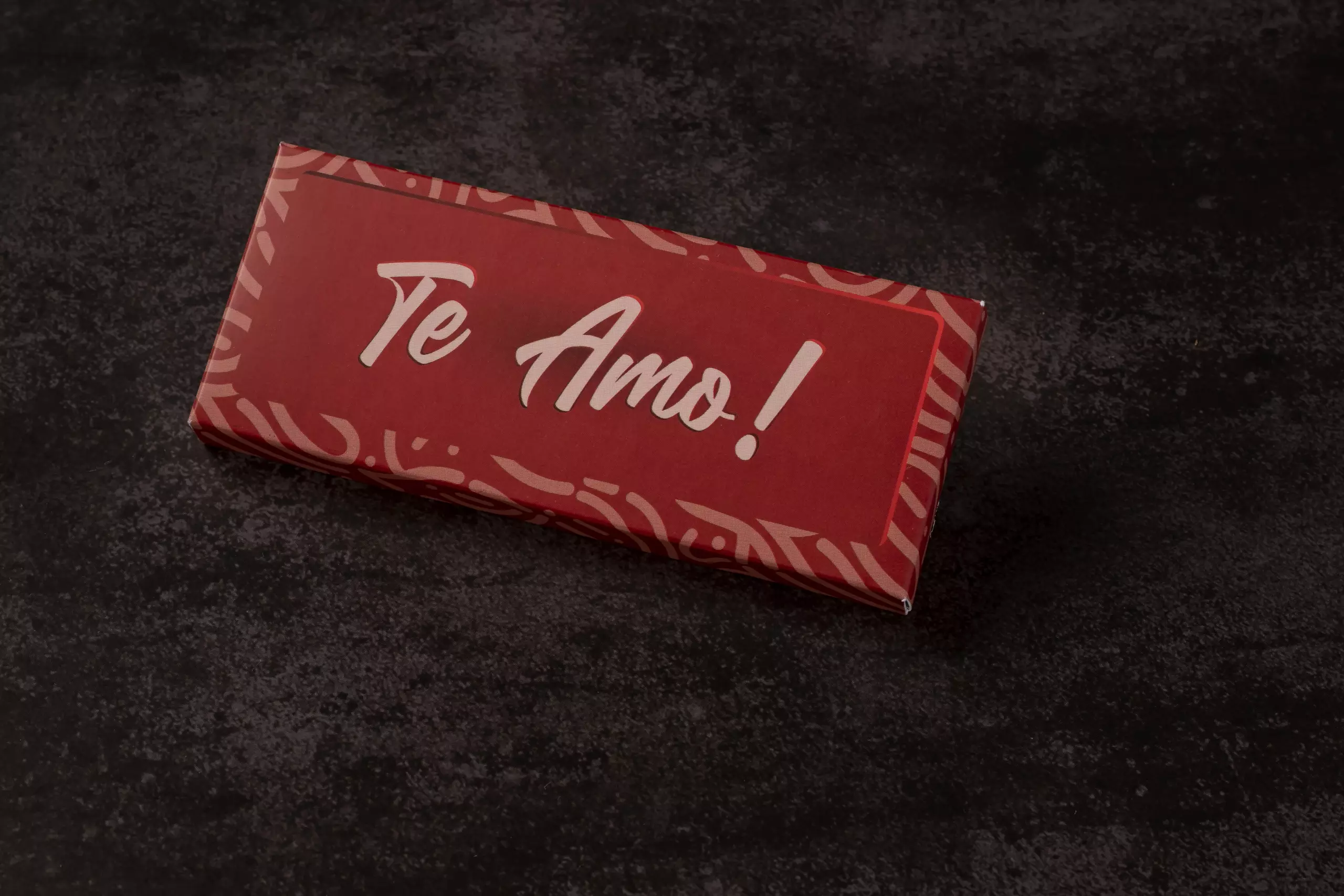 Uma barra de chocolate retangular com uma embalagem de papel vermelha. A mensagem "Te Amo!" está escrita em branco no centro, com uma fonte grande e estilizada. A barra está disposta em ângulo sobre uma superfície escura e texturizada.