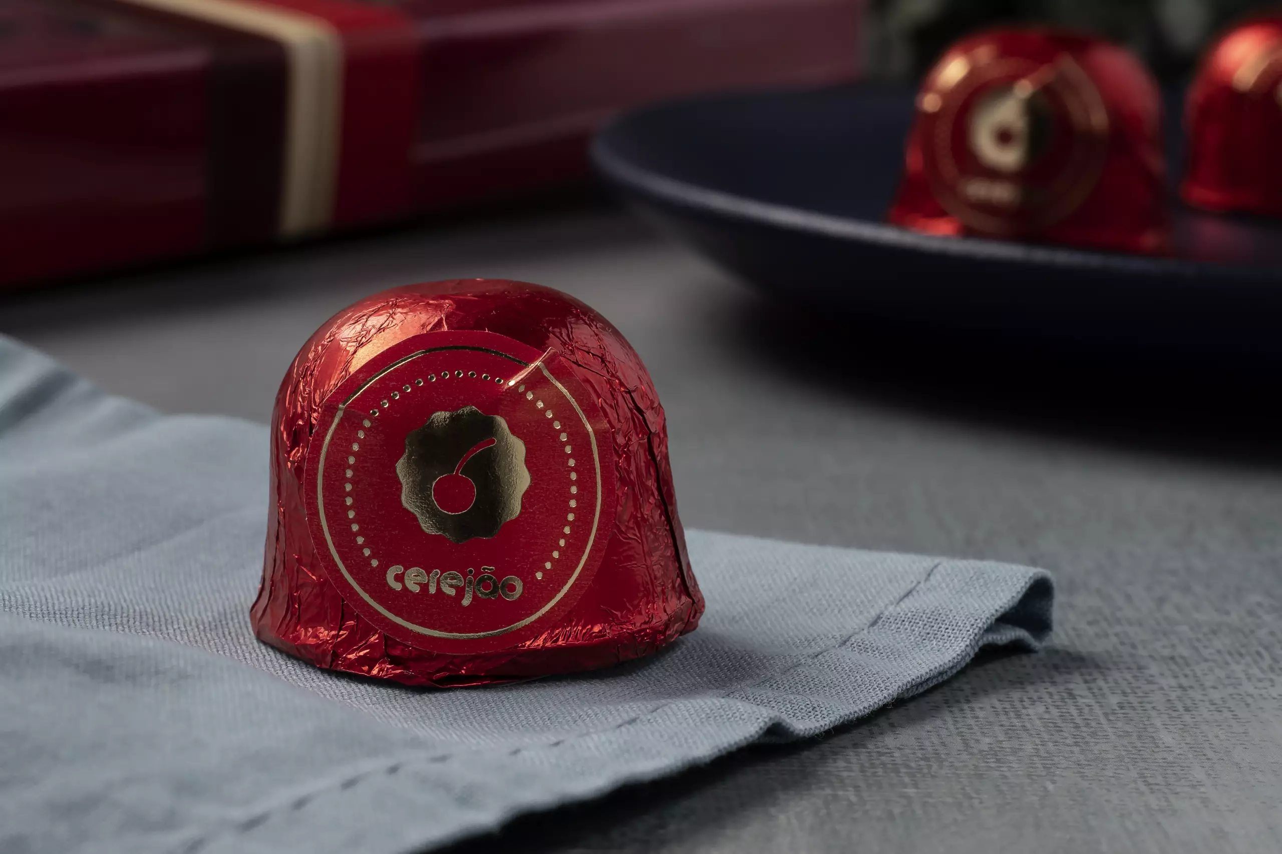 Close-up de um bombom de chocolate de cereja embalado em papel laminado vermelho brilhante. No topo, um selo circular dourado exibe a palavra "Cerejão" e o desenho de uma cereja. O bombom está sobre um guardanapo de tecido cinza-azulado, com um prato escuro e uma caixa de presente desfocados ao fundo. Do lado direito, tem mais bombons embalados emcima de um prato.