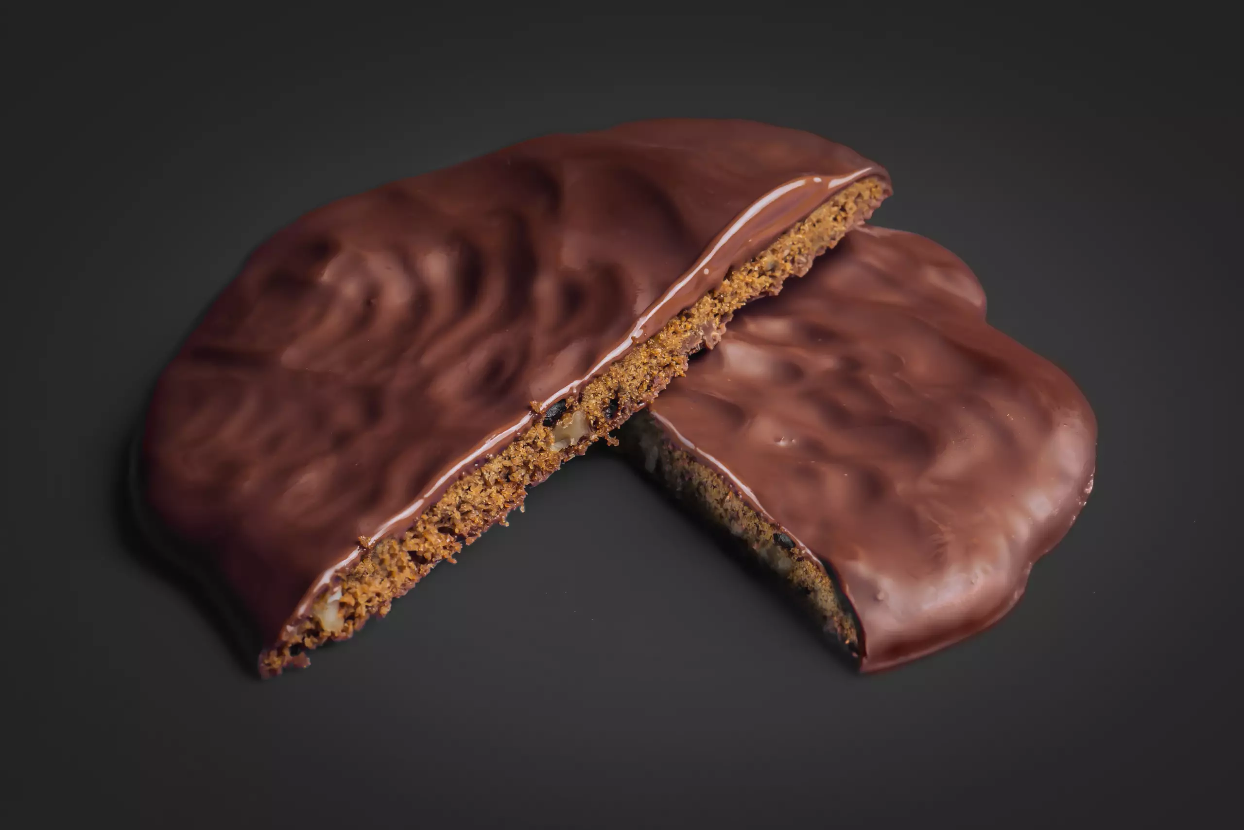 Close-up de um cookies artesanal banhados em chocolate ao leite, mostrando o interior com a massa dourada com pedaços de nozes e chocolate, sobre fundo preto.