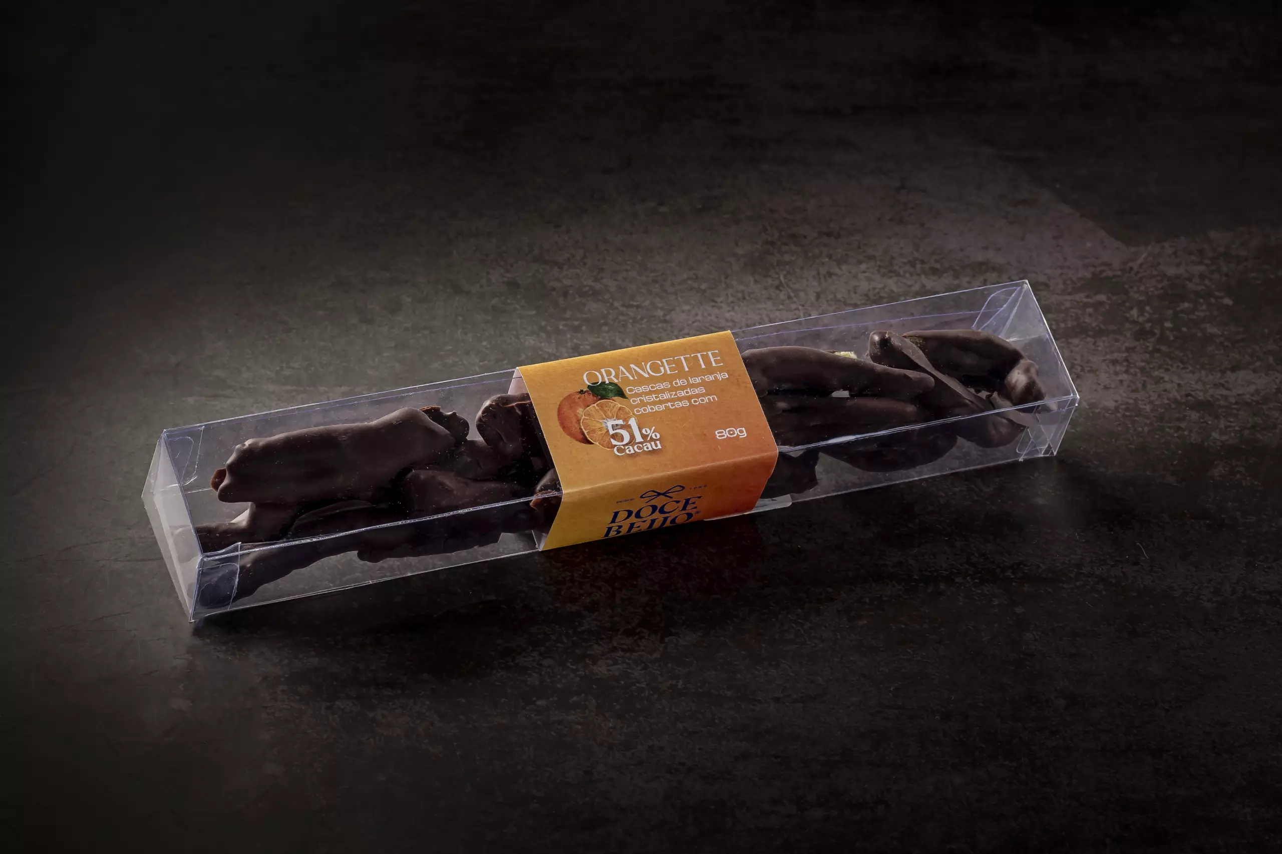 Uma caixa retangular de acetato transparente, com Orangettes (tiras de laranja cristalizadas cobertas com chocolate). A embalagem é selada por um rótulo de papel que informa o nome do produto, os ingredientes "51% cacau" e a marca "Doce Beijo". O produto está sobre uma superfície escura e texturizada.