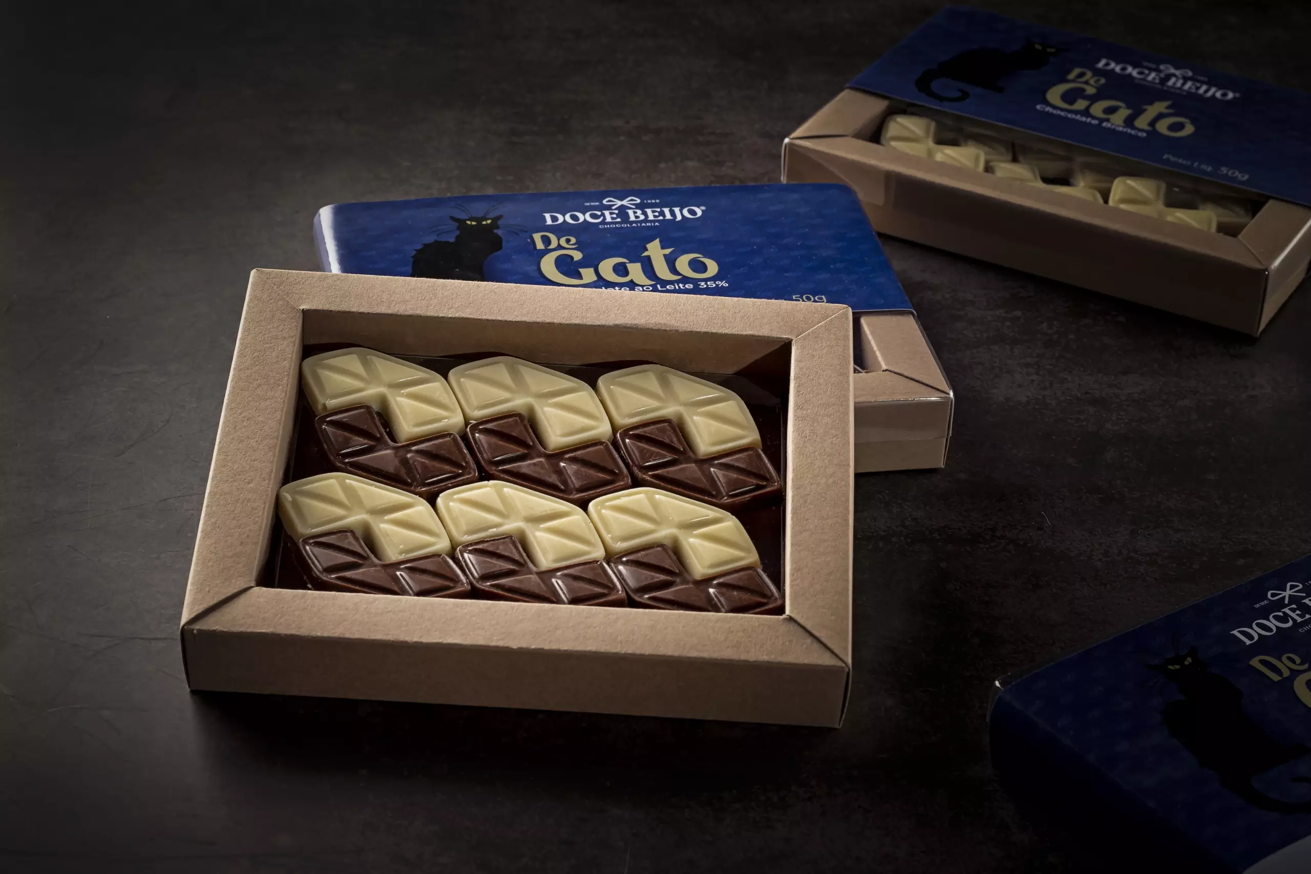 Três caixas do chocolate "Beijo de Gato" em uma superfície escura. A caixa em primeiro plano está aberta, exibindo seis chocolates retangulares com um padrão geométrico. Cada chocolate é bicolor, com metade de chocolate ao leite e metade de chocolate branco, dispostos de forma organizada.
