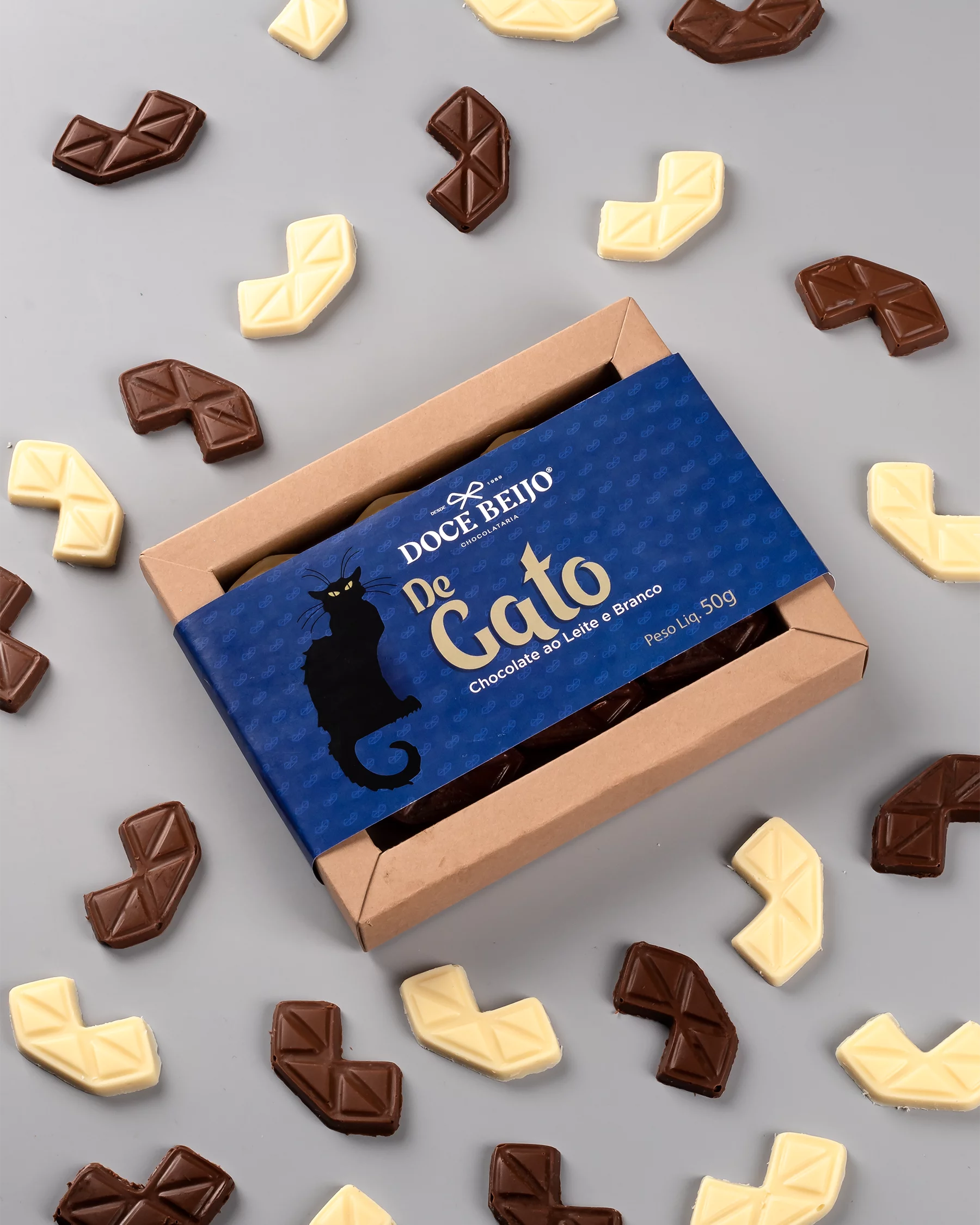 Uma caixa de papel kraft aberta do chocolate "Do Gato", da marca "Doce Beijo". A embalagem possui uma cinta azul com a silhueta de um gato preto. Vários pequenos chocolates, nos sabores ao leite e branco, estão dentro da caixa e espalhados ao redor sobre um fundo cinza.