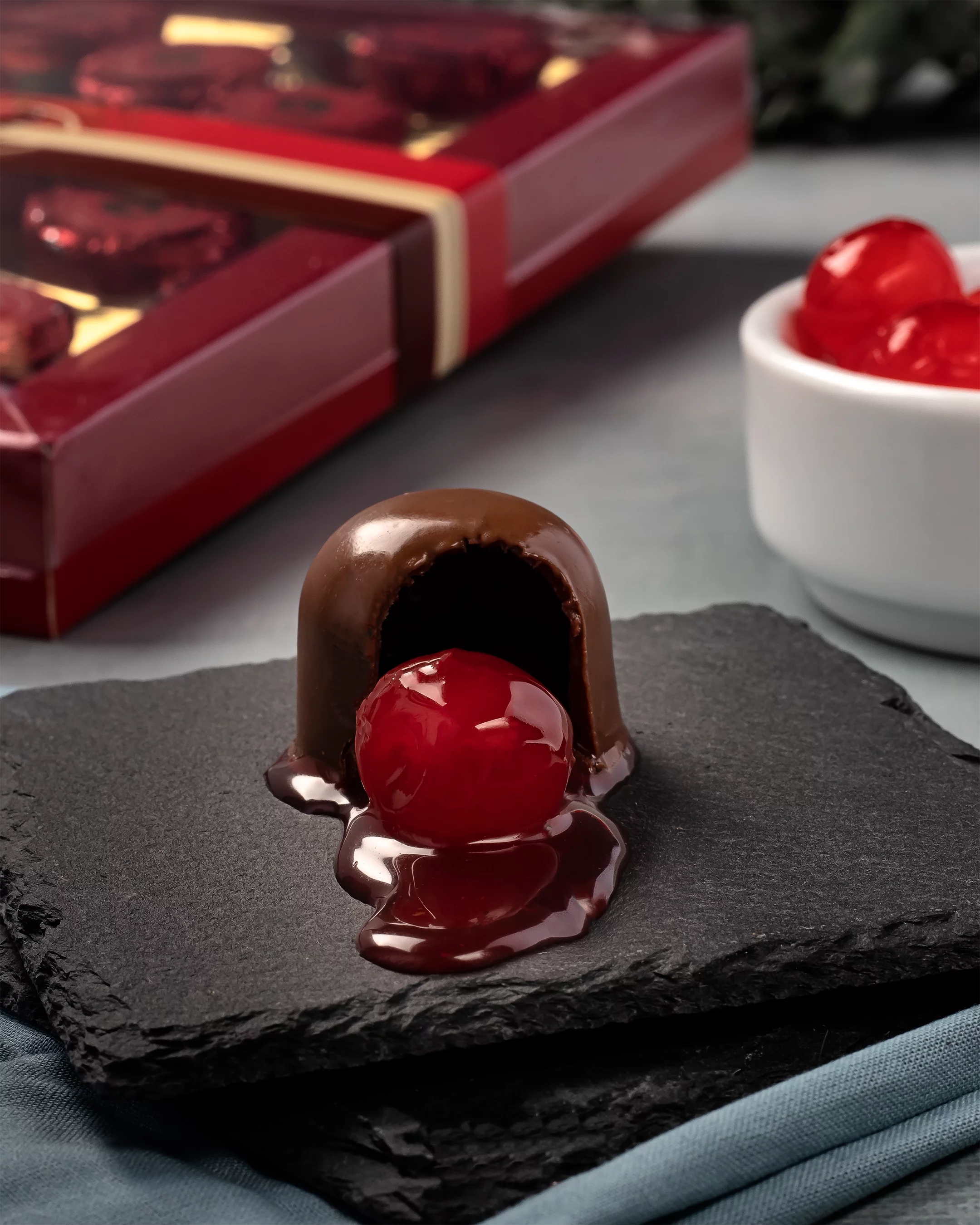 Um bombom de chocolate em formato de cúpula em close-up, posicionado sobre uma placa de ardósia preta. O bombom está aberto, revelando uma cereja vermelha e brilhante em calda saindo de seu interior. Ao fundo, um pote branco com mais cerejas e uma caixa de presente vermelha com bombons completam a cena.
