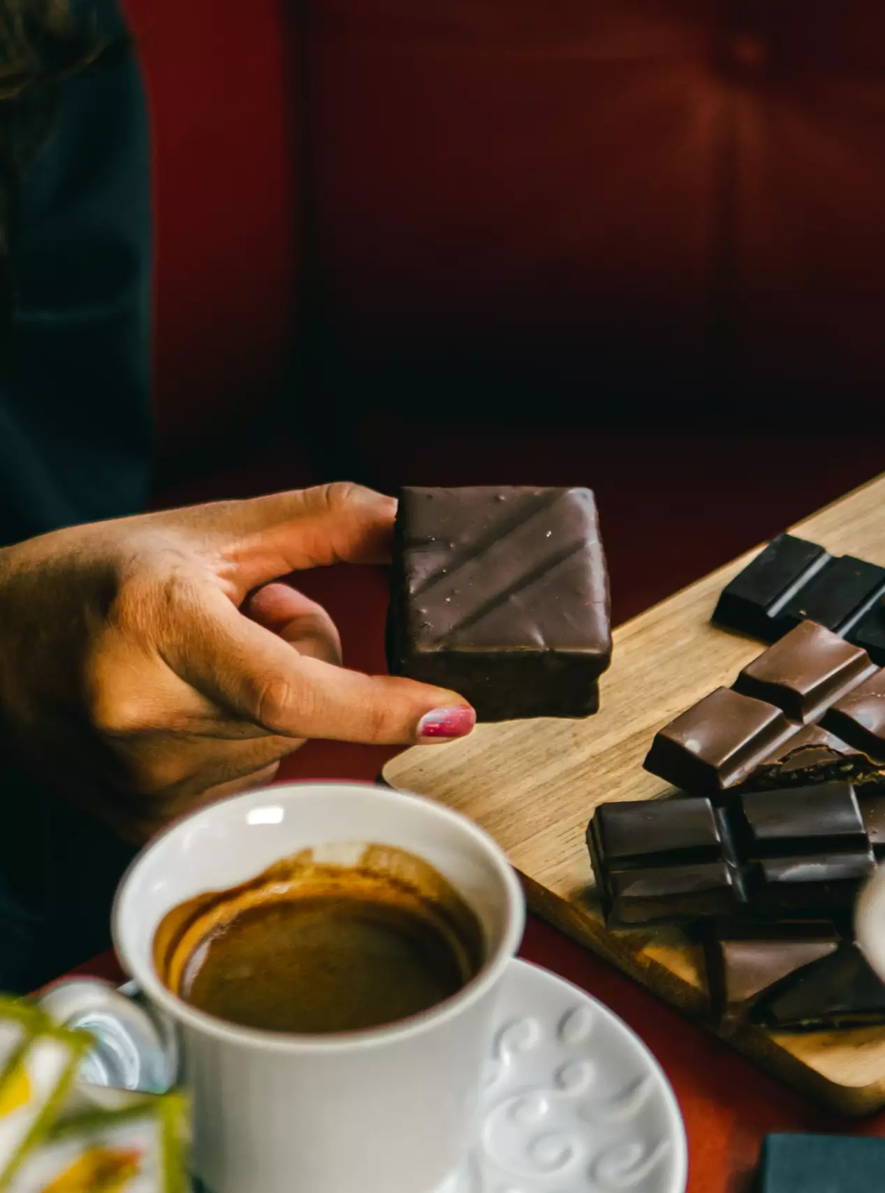 A mão de uma pessoa segura um chocolate quadrado com decoração de linhas diagonais. Embaixo, uma tábua de madeira está repleta de outros chocolates quadrados, e em primeiro plano, uma xícara branca com café preto. O fundo vermelho escuro.