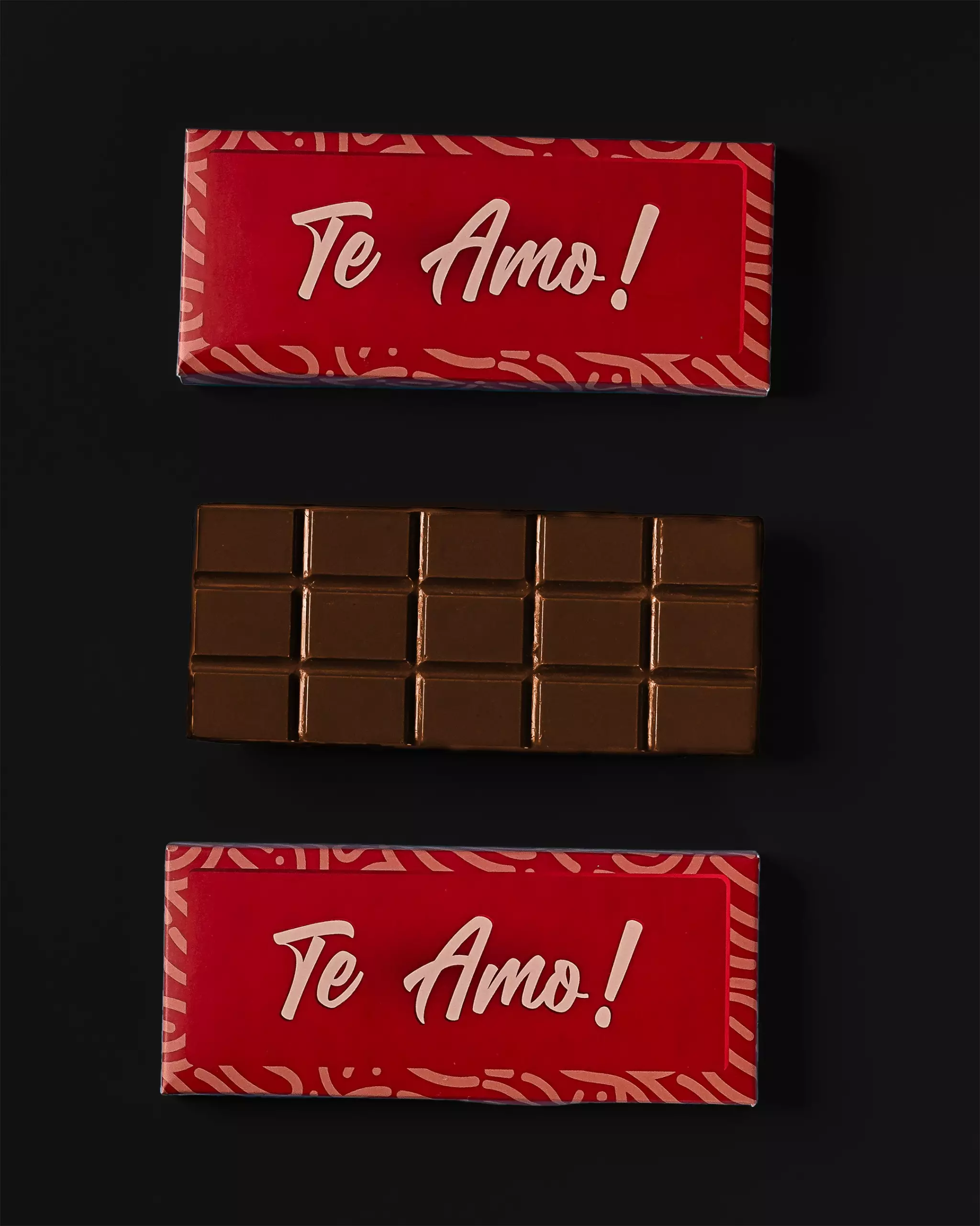 Composição com uma barra de chocolate ao leite e sua embalagem. A embalagem vermelha com a mensagem "Te Amo!" em branco aparece acima e abaixo da barra de chocolate, que está desembalada no centro, sobre um fundo preto.