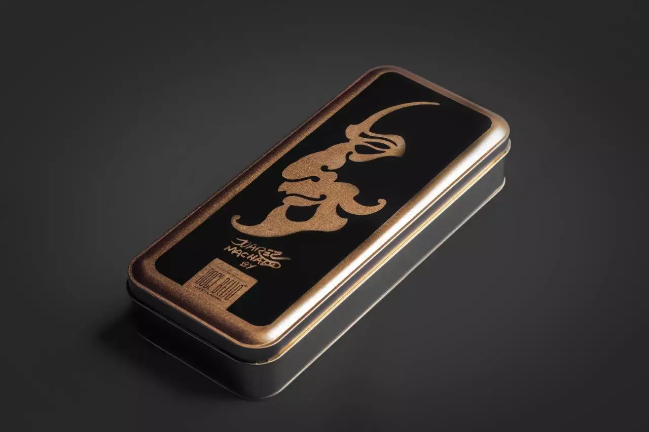 Lata de chocolate retangular de metal, predominantemente preta com detalhes e bordas em dourado, vista em perspectiva sobre um fundo cinza-escuro. A tampa da lata apresenta uma ilustração estilizada em dourado do perfil de um rosto, acompanhada da assinatura "Juarez Machado '87" e o logotipo da "Doce Beijo".