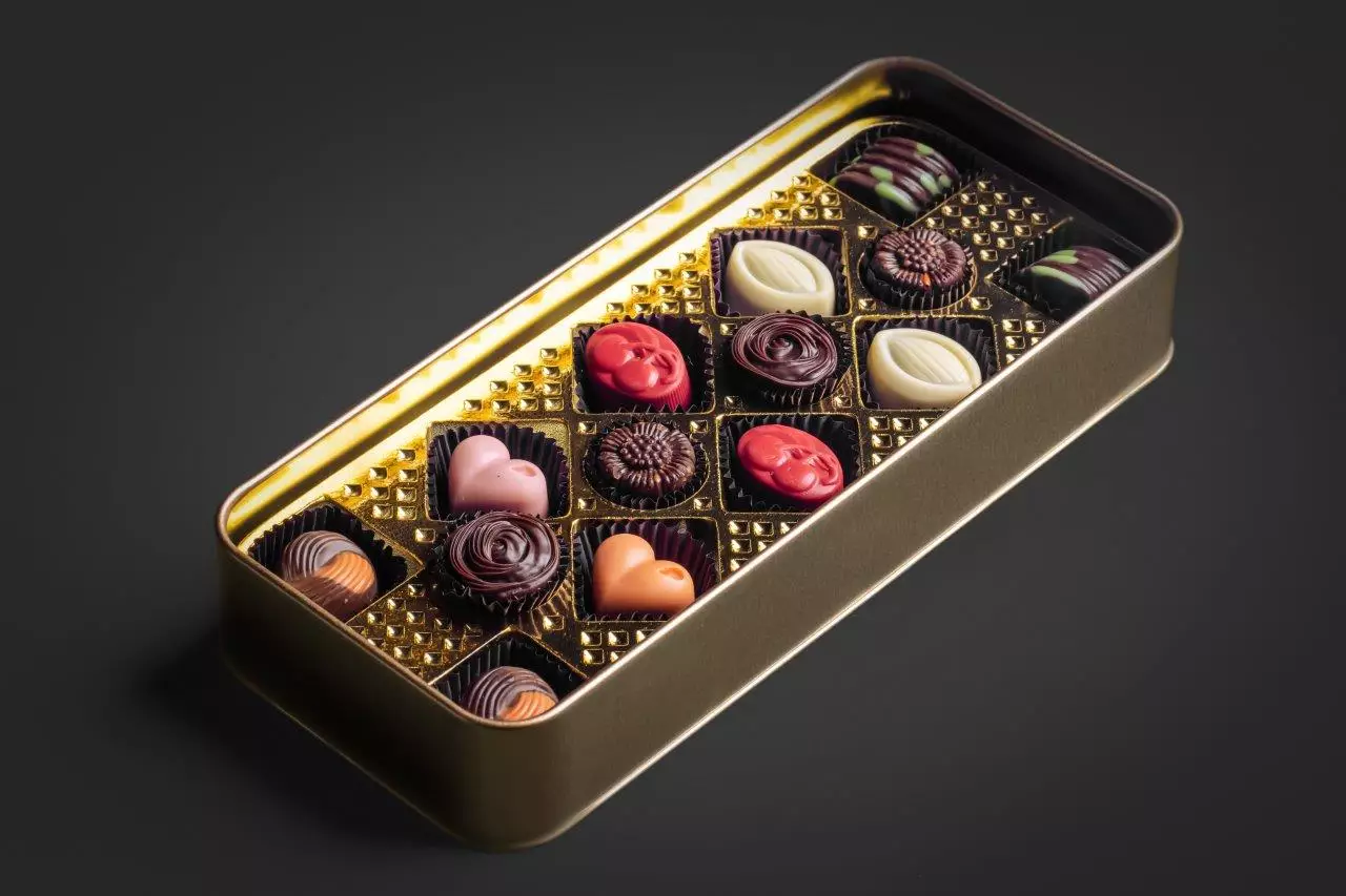Vista de cima de uma lata de bombons aberta. Os bombons possuem diversos formatos, como coração, flor e folha, e diferentes coberturas e cores, organizados em uma bandeja interna dourada.