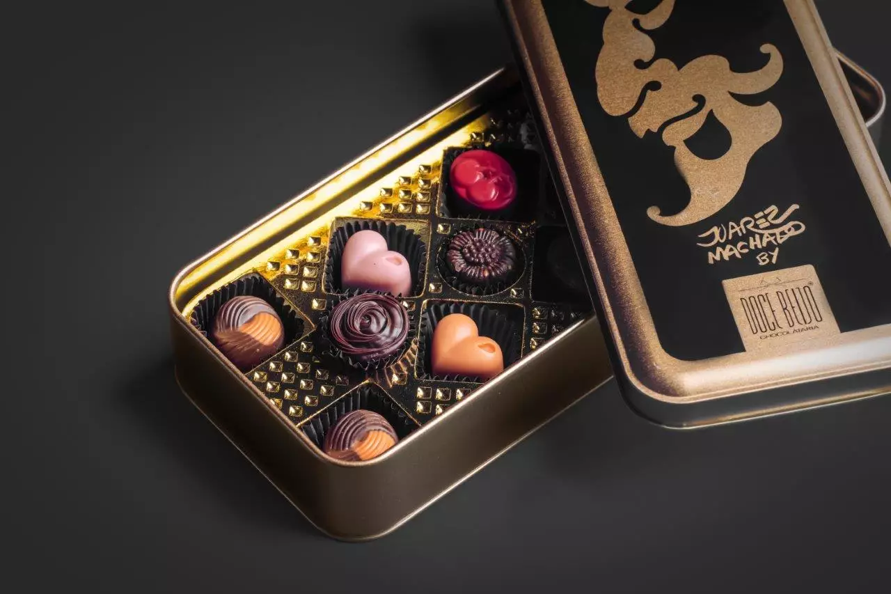 Uma lata de metal preta e dourada, com a tampa aberta exibindo a arte de Juarez Machado. Dentro, uma variedade de bombons artesanais de diferentes formatos (coração, flor) e cores está disposta em uma bandeja dourada.